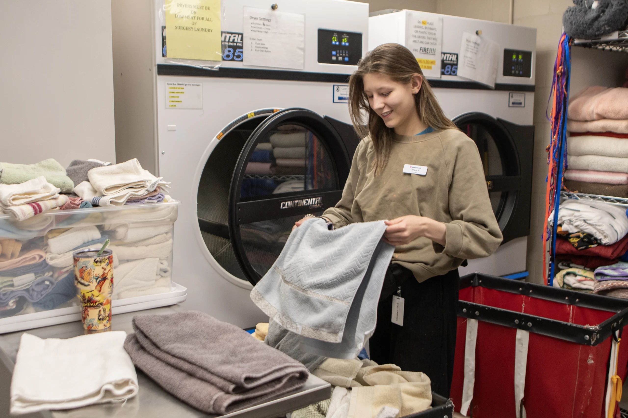 Young woman in olive sweatshirt folding gray towel in veterinary laundry room with industrial dryers and stacked linens