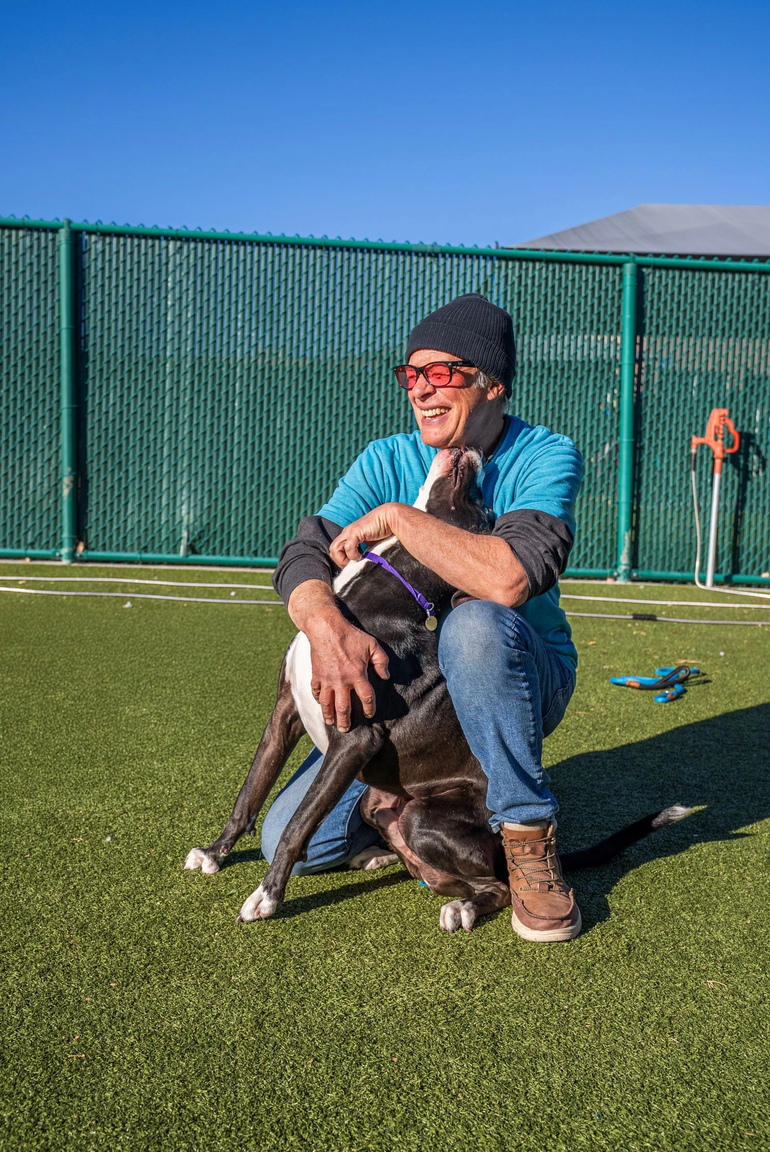 Smiling man in beanie and sunglasses crouches on turf while a gray and white pit bull leans affectionately against him
