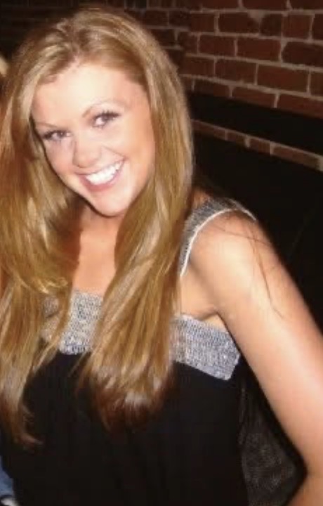 Young woman with long blonde hair smiling, wearing a black top with silver metallic trim, posed against a brick wall