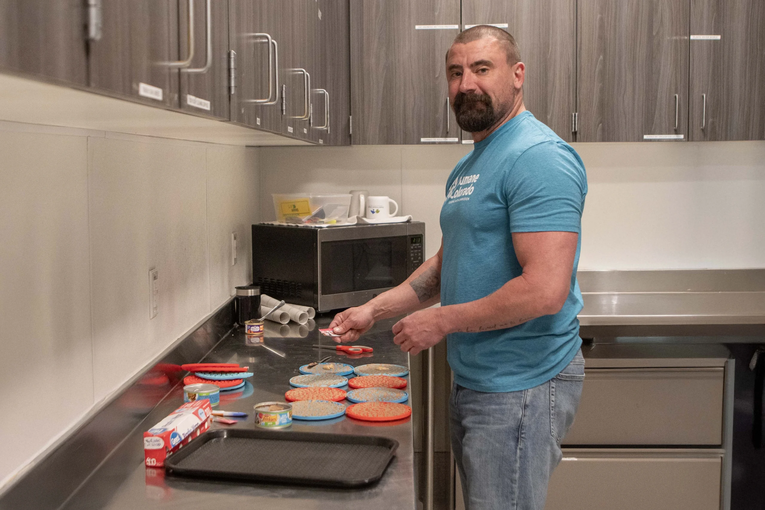 Volunteer in Humane Colorado shirt prepares food on small plates in shelter kitchen, with canned food and supplies nearby