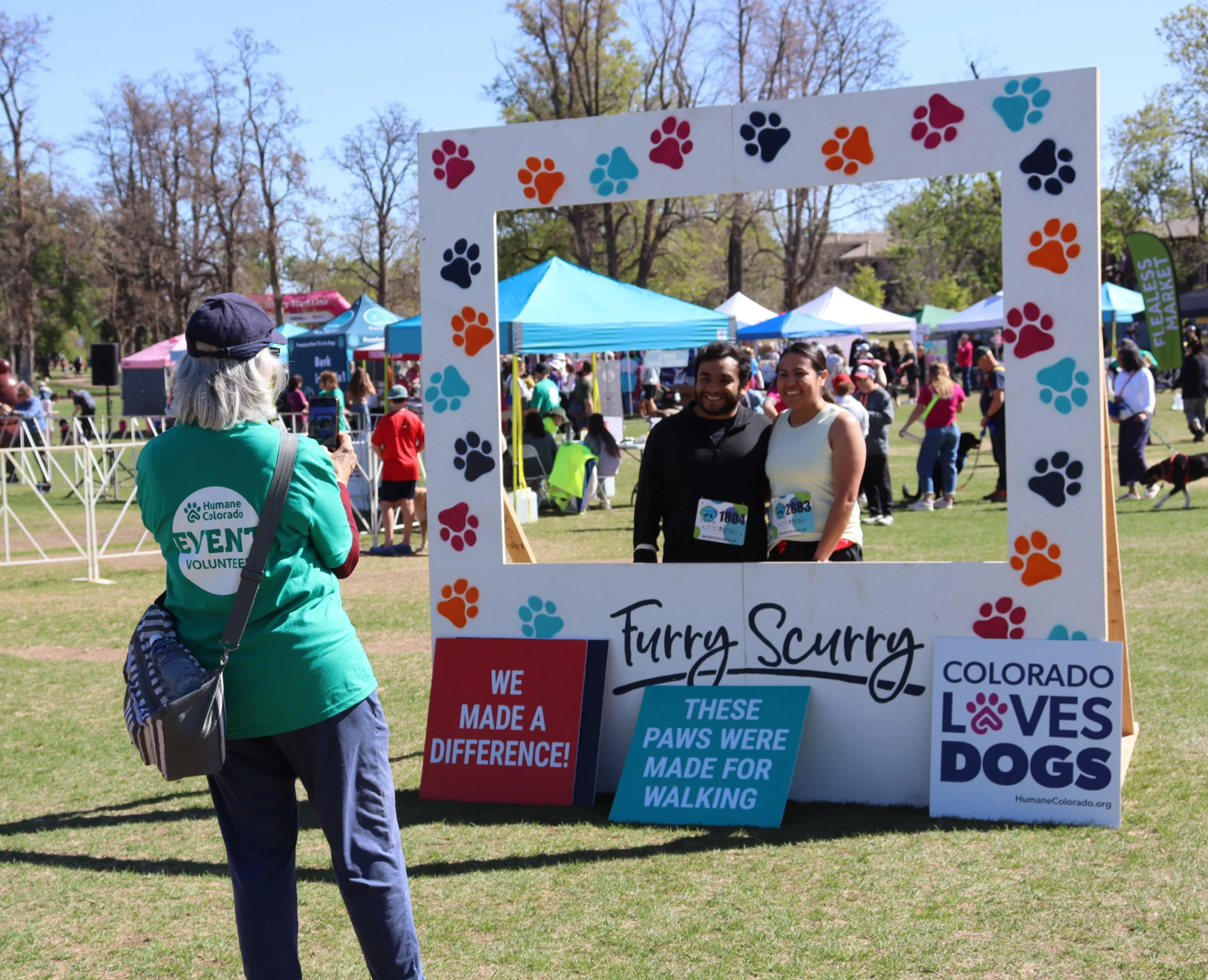 Volunteer photographs couple posing in Furry Scurry photo frame decorated with paw prints at outdoor Humane Colorado event
